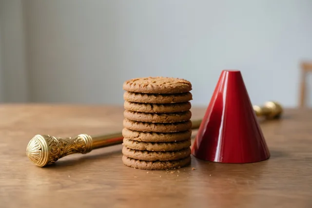Een stapel ronde bruine speculaaskoeken staat op een houten tafel naast een rode mijter en een gouden staf.