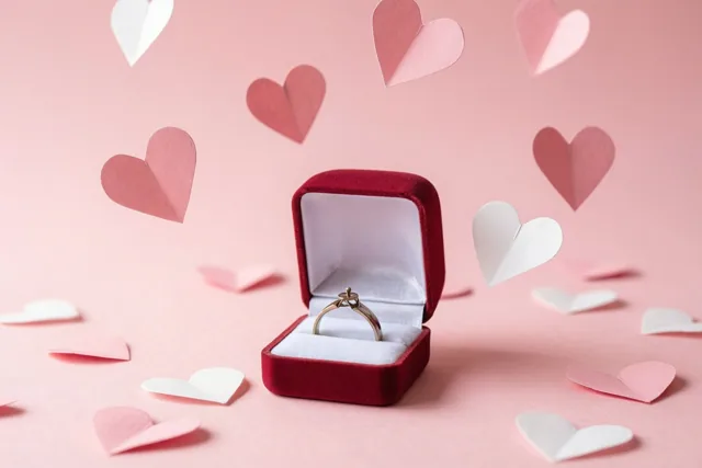 Een open rood fluwelen doosje met een gouden ring staat op een zachtroze ondergrond, met vallende papieren hartjes in roze en wit errond.