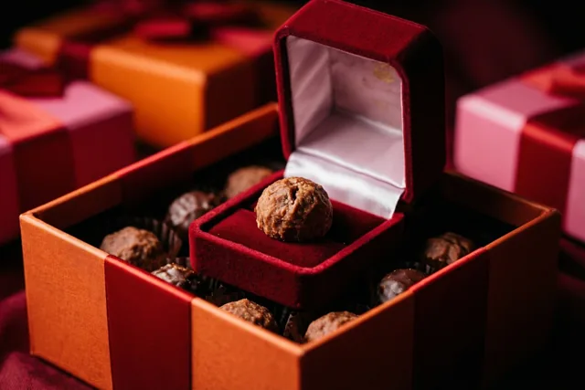 Een klein open rood fluwelen ringdoosje staat in een grotere geschenkdoos met Belgische pralines, tegen een donkere achtergrond met wazige geschenkdozen.