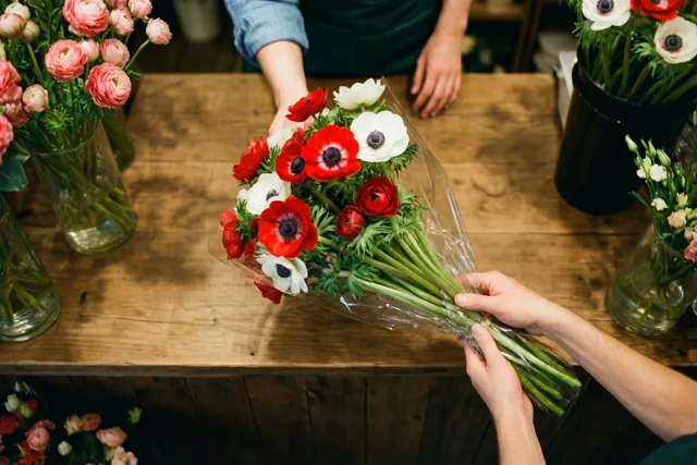 Een ingepakt boeket rode en witte anemonen wordt over een houten toonbank in een bloemenwinkel doorgegeven, met andere bloemstukken op de achtergrond.