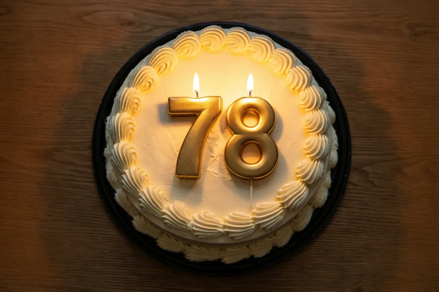 Een ronde roomtaart met gespoten versiering en twee brandende gouden cijferkaarsen die 78 vormen, staat op een houten tafel.