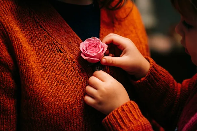 Kleine kinderhanden steken een roze roos in het knoopsgat van de oranje gebreide vest van een moeder, in warm licht.