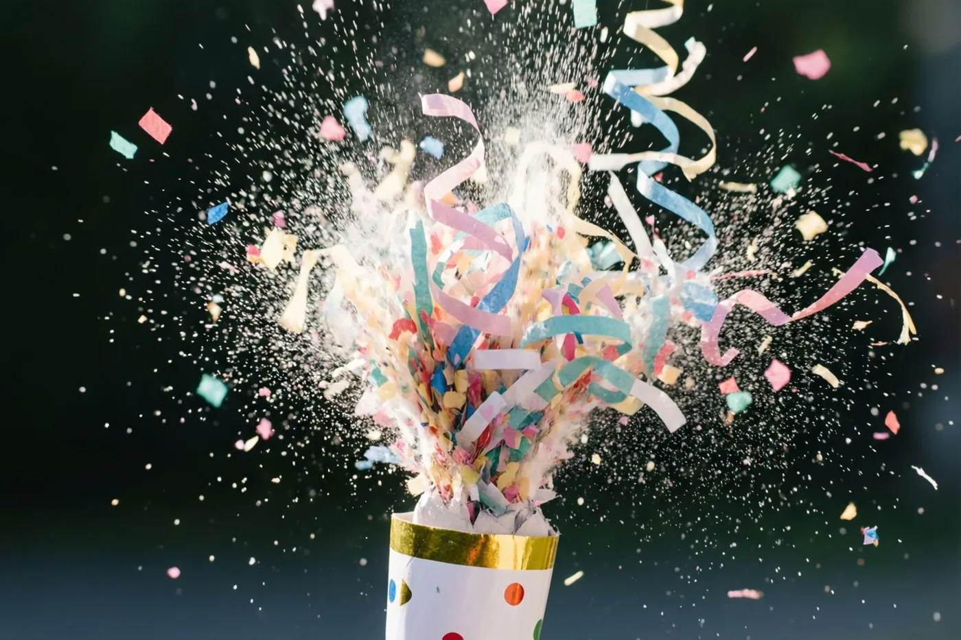Een knalbonbon barst open met confetti en gekruld papier in roze, blauw en goud tegen een donkere achtergrond.