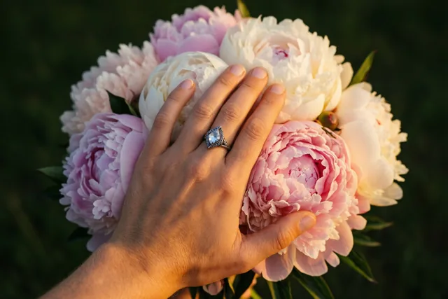 Een hand met een ring met één steen aan de ringvinger houdt een boeket roze en witte pioenrozen vast in warm goudkleurig licht tegen een onscherpe groene achtergrond.