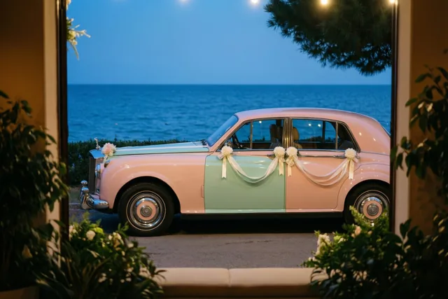 Door een raam met groen op de voorgrond is een roze en mintgroene oldtimer met witte linten en bloemen te zien aan zee, met kalm blauw water en een schemerlucht erachter.