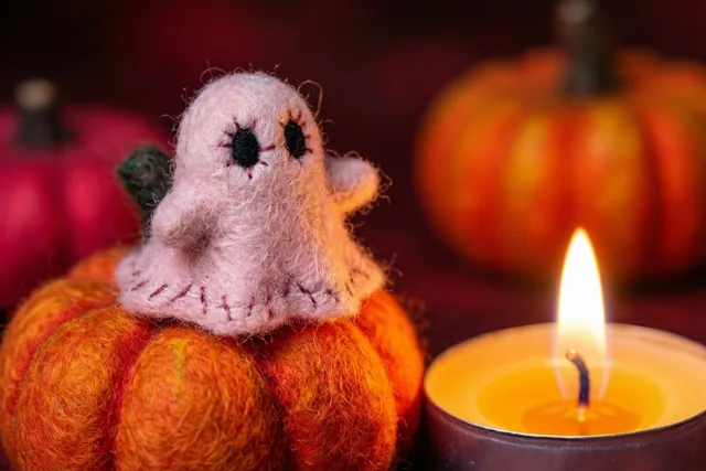 Een klein handgemaakt roze vilten spookje met geborduurde ogen zit op een oranje vilten pompoen naast een brandend theelichtje, met andere pompoenen op de donkere wazige achtergrond.