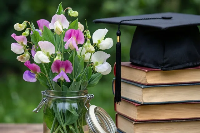 Een glazen pot met roze, witte en paarse reukerwten staat naast oude boeken met daarop een zwarte afstudeerhoed, met wazig groen op de achtergrond.