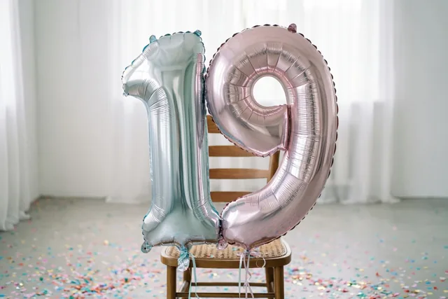 Een zilveren en een pastelroze folieballon in de vorm van 1 en 9 zijn vastgebonden aan een houten stoel in een lichte kamer met confetti op de vloer.