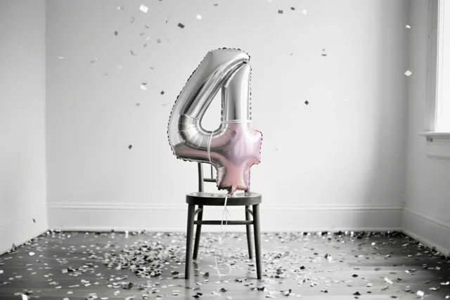 Een zilveren en pastelroze cijferballon 4 hangt aan een houten stoel in een lichte kamer, met zilveren confetti in de lucht en op de vloer.
