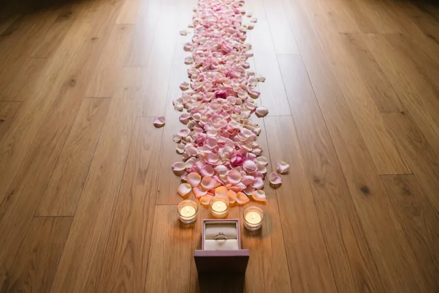 Een pad van roze rozenblaadjes op een houten vloer leidt naar een open ringdoosje met drie brandende kaarsen eromheen.