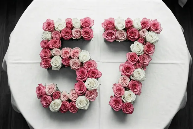 Van bovenaf is het getal 57 te zien, gelegd met roze en witte rozen op een wit tafelkleed in zachte tinten.