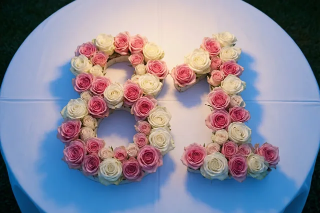 Het getal 81 van roze en witte rozen ligt op een wit tafelkleed buiten op het gras, van bovenaf gezien in zacht licht.