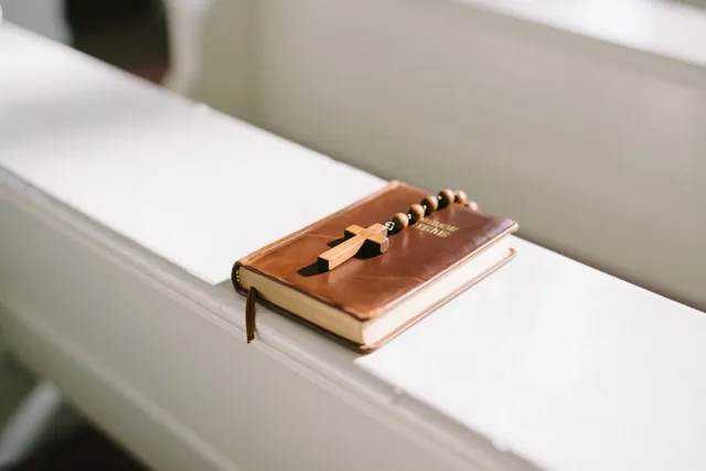 Een houten rozenkrans ligt op een gesloten bruinleren gebedenboek op een witte kerkbank in zacht licht.