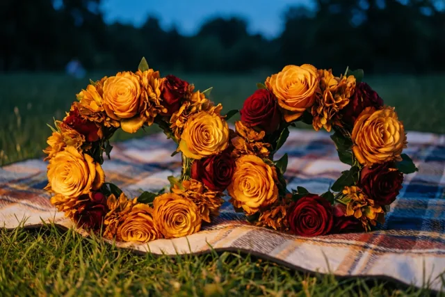 Twee bloemenkronen van gele en rode rozen liggen op een geruit picknickdeken op het gras, met een donkerblauwe avondlucht en onscherpe bomen op de achtergrond.