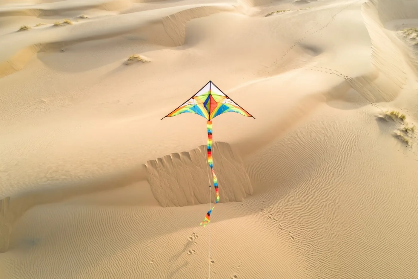 Een kleurrijke ruitvormige vlieger met een regenboogstaart vliegt laag boven goudkleurige zandduinen in warm zonlicht.