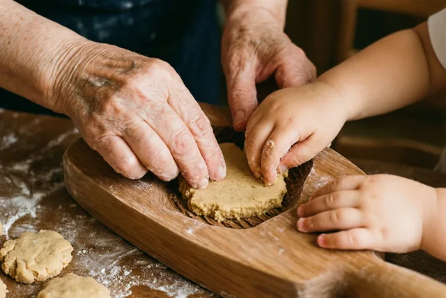 De handen van een oudere persoon en een kind drukken deeg in een houten vorm op een met bloem bestoven houten werkblad.
