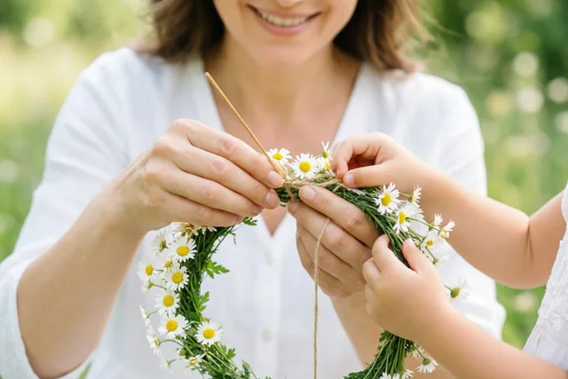 De handen van een moeder en een kind vlechten buiten samen een kroon van witte madeliefjes met gele hartjes en groene stelen.