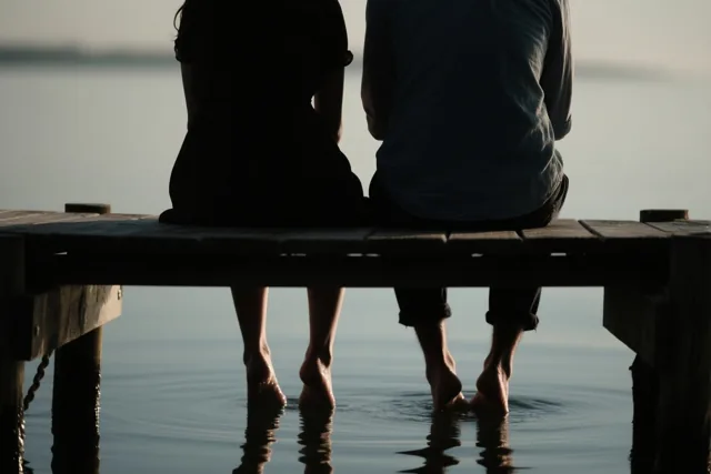 Twee mensen zitten met hun rug naar de kijker op een houten steiger met hun blote voeten in rustig water, in zachte blauwe en grijze tinten.