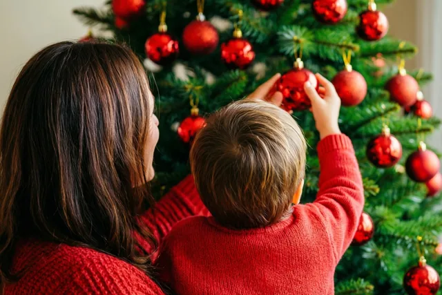 Een ouder en een klein kind hangen samen rode kerstballen in een kerstboom, van achteren gezien in een beeld met vooral rood en groen.