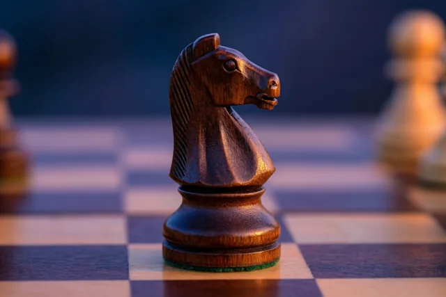 Een houten schaakpaard staat op een schaakbord in warm goudkleurig licht, met onscherpe schaakstukken en een donkerblauwe achtergrond.