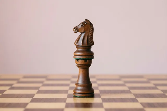 Een houten schaakpaard staat rechtop op een houten schaakbord tegen een effen zachtroze achtergrond.