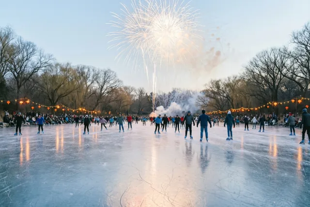 Schaatsers bewegen over een bevroren vijver tussen kale bomen, met lantaarnsnoeren langs de randen en vuurwerk in de schemerlucht.