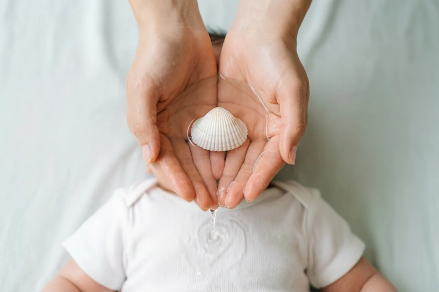 Twee handen van een volwassene houden een kleine witte schelp met water boven het hoofd van een baby in een wit kleed, tegen een zachte witte achtergrond.