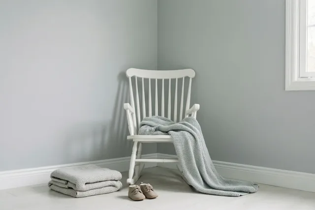 Over een witte houten schommelstoel in een lichtgrijze babykamer ligt een lichtgrijs gebreid dekentje, met een opgevouwen bijpassend dekentje en kleine beige schoentjes op de lichte houten vloer.
