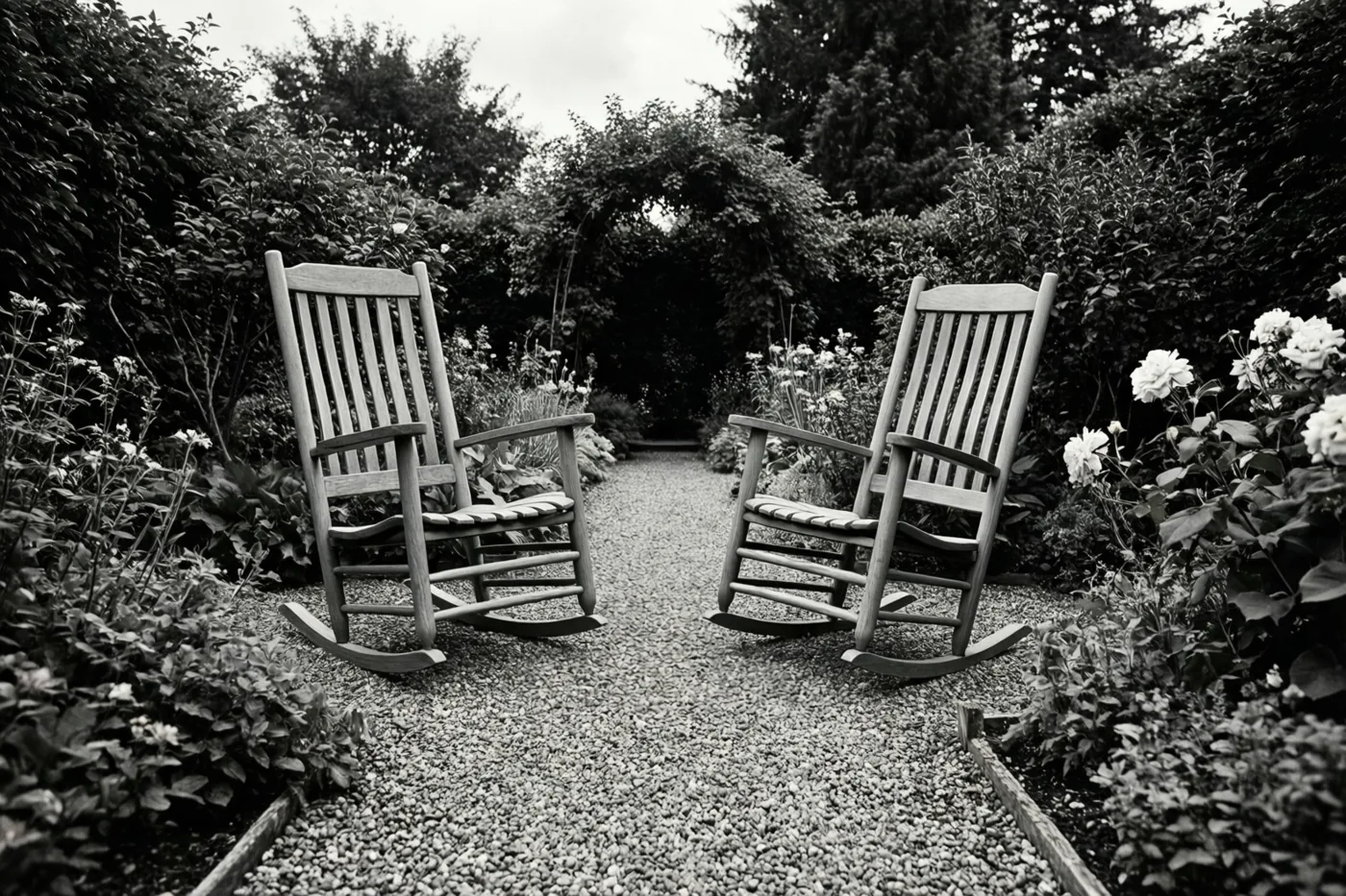 Een zwart-witbeeld toont twee houten schommelstoelen tegenover elkaar op een grindpad in een weelderige tuin met dicht gebladerte en bloeiende struiken.