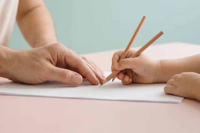 Een volwassen hand begeleidt een kinderhand met een potlood boven een wit blad op een roze oppervlak, tegen een lichtblauwe achtergrond.
