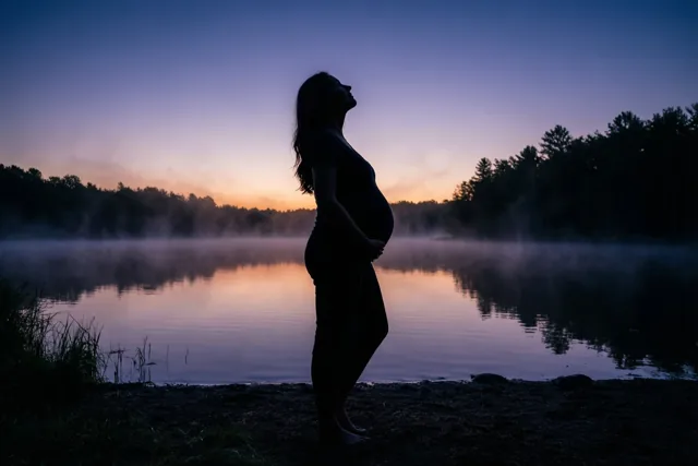 Een zwangere vrouw staat als silhouet met haar hoofd omhoog aan een kalm meer bij dageraad, met mist boven het water en een weerspiegeling van de diepblauwe tot zachtoranje lucht.