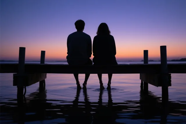 Twee mensen zitten als silhouetten naast elkaar op een houten steiger met hun voeten in kalm water onder een paarse en oranje avondlucht.