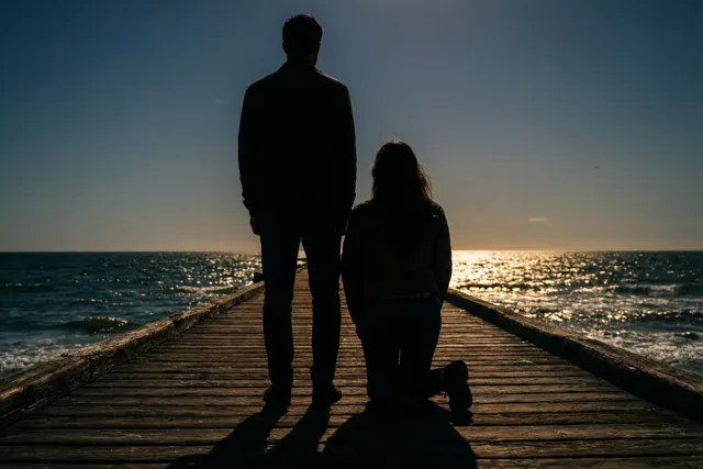 Twee silhouetten zijn van achteren te zien op een houten pier, één staand en één knielend, met diepblauw water en gouden weerspiegelingen van de ondergaande zon.