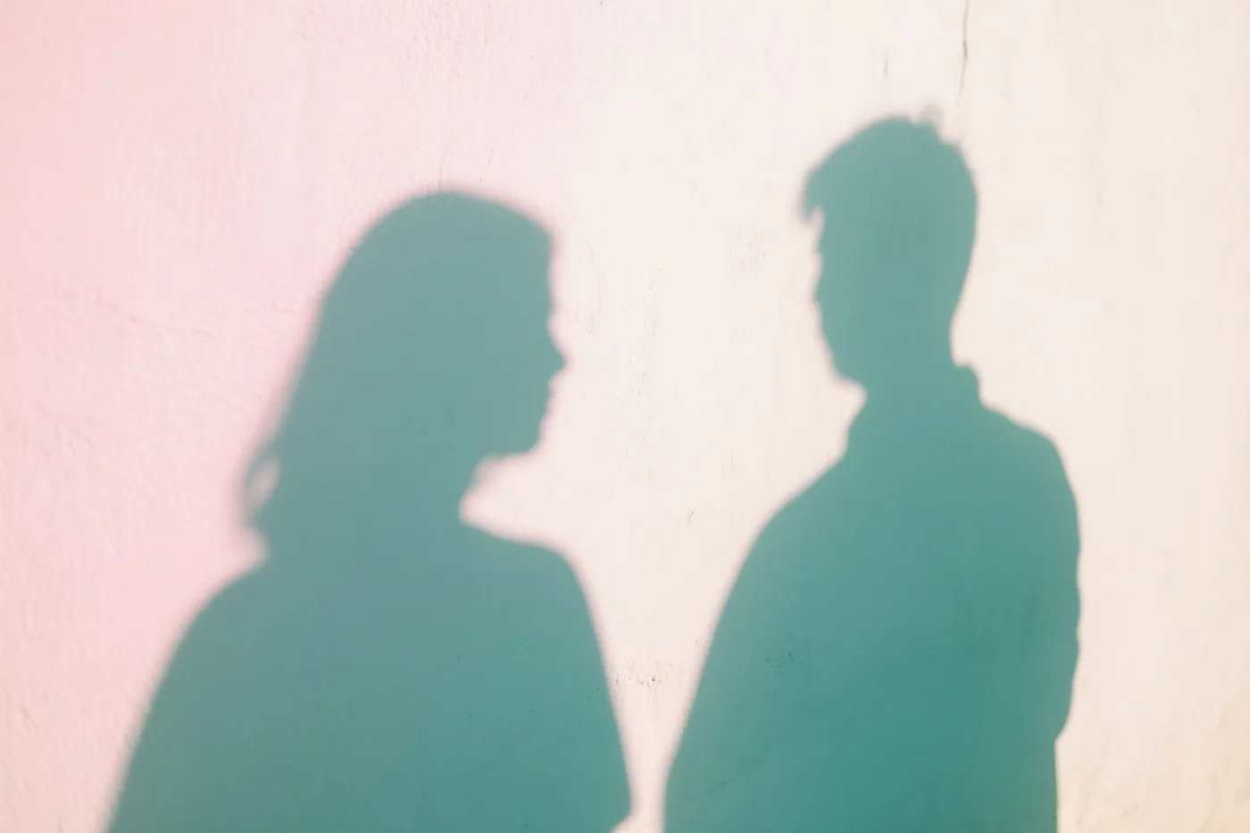 Twee silhouetten van mensen die rug aan rug staan, werpen overlappende schaduwen op een witgekalkte muur in zachtroze, gebroken witte en blauwgroene tinten.