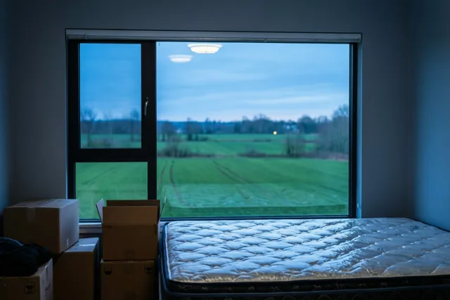 Een matras en kartonnen verhuisdozen liggen voor het raam van een kale slaapkamer, met uitzicht op groene velden onder een schemerblauwe lucht.