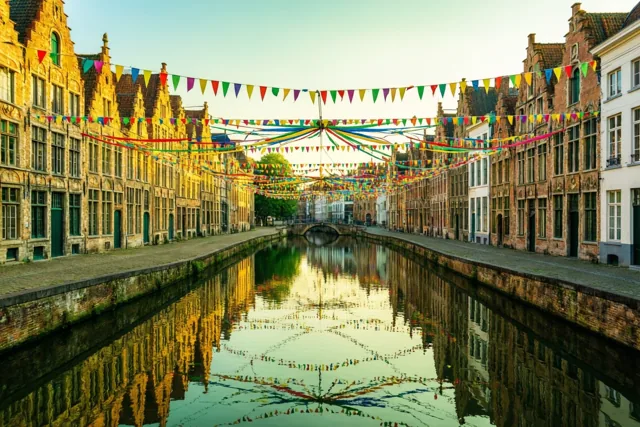 Carnavalsvlaggetjes en slingers hangen boven een kanaal tussen oude Vlaamse gebouwen en weerspiegelen in het kalme water in warm gouden licht.