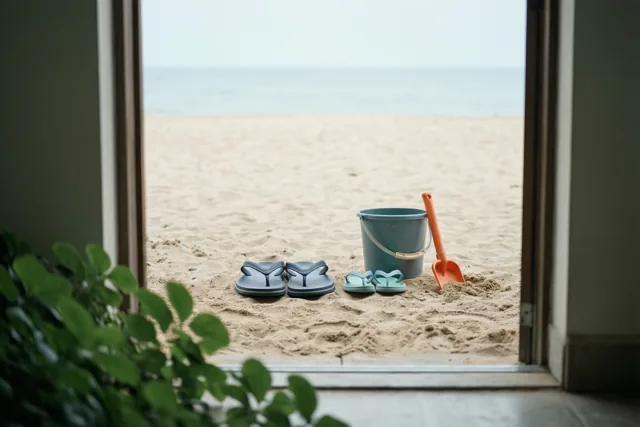 Door een deuropening zijn grote en kleine teenslippers op een zandstrand te zien naast een blauwe emmer en een oranje schopje.