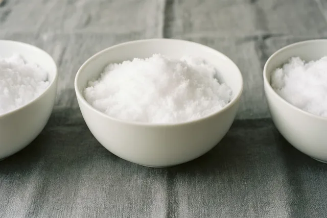 Drie witte porseleinen schaaltjes gevuld met sneeuw staan op grijs linnen in een sober stilleven met witte en grijze tinten.