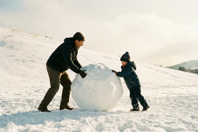 Een vader en een kind duwen een grote sneeuwbal een besneeuwde heuvel op in een wit landschap met zacht, warm licht.