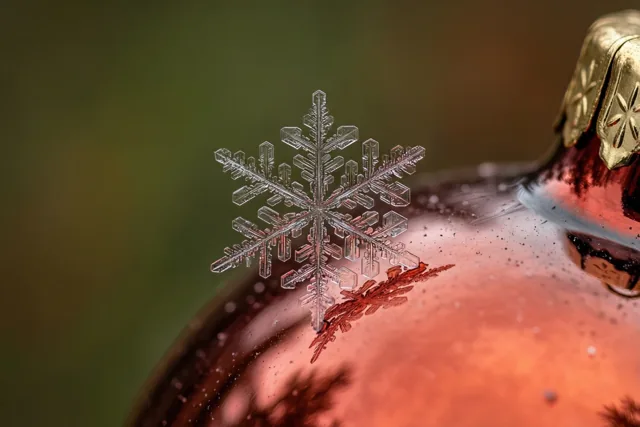 Een fijne sneeuwvlok rust op de ronde kant van een glanzende rode glazen kerstbal, tegen een wazige achtergrond in groene en bruine tinten.