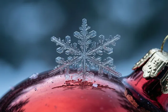 Een fijne sneeuwvlok ligt op de gebogen kant van een glanzende rode glazen kerstbal, tegen een wazige blauwgrijze achtergrond.