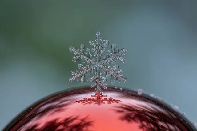 Een fijne sneeuwvlok ligt op een glanzende rode glazen kerstbal, tegen een zacht vervaagde achtergrond in groen en blauw.