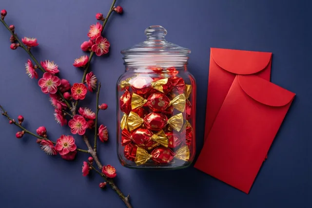 Een glazen pot met snoepjes in rode en goudkleurige wikkels staat naast twee rode enveloppen en een tak met roze pruimenbloesems tegen een donkerblauwe achtergrond.