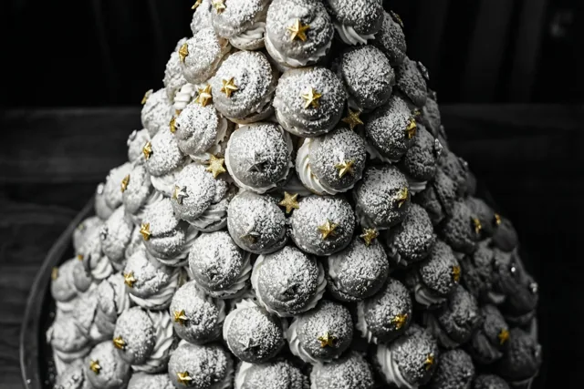 Een hoge kegelvormige toren van roomsoesjes met poedersuiker en kleine gouden sterren staat op een donkere ondergrond tegen een zwarte achtergrond.