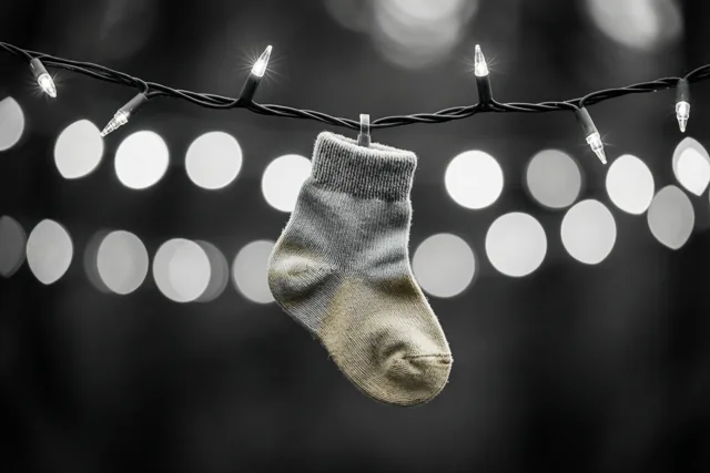 Een gedragen babysokje hangt aan een brandend lichtsnoer voor een donkere achtergrond met wazige lichtcirkels op een zwart-witfoto.