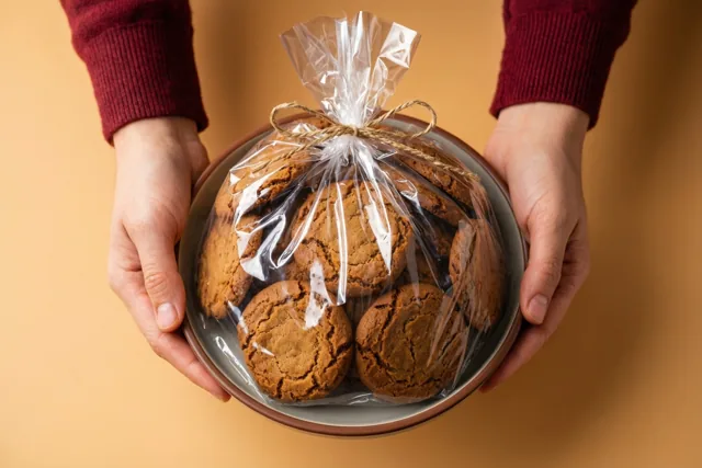 Twee handen uit een bordeauxrode trui houden een bord zelfgebakken speculoos in doorzichtig cellofaan met een touwstrik vast tegen een effen warmbeige achtergrond.