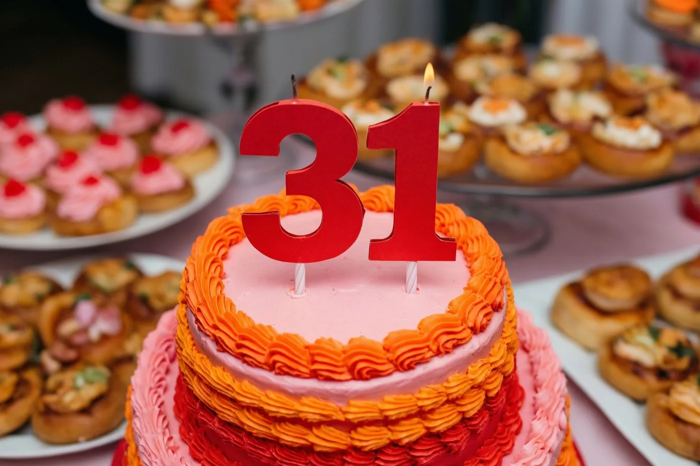 Een stapeltaart met roze en oranje glazuur, een rode papieren 31 en één brandend kaarsje staat tussen borden met gebak op een buffet.