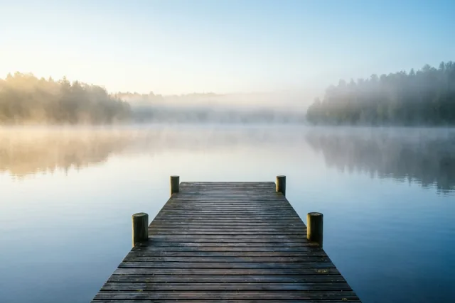 Een houten steiger loopt een kalm meer met zachte ochtendmist in, met bomen in de verte onder een heldere hemel.