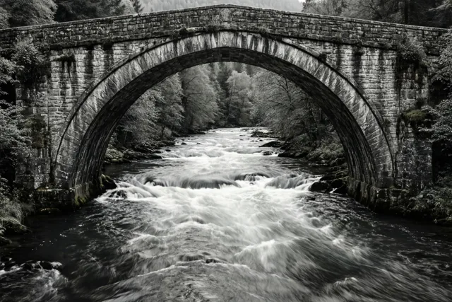 Een zwart-witfoto toont een stenen boogbrug over een snelstromende rivier in een bosrijke omgeving.