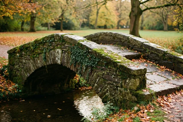 Een oude stenen voetbrug met mos en klimop overspant een kalme beek in een herfstpark, tussen oranjebruine bladeren en bomen met gouden loof.